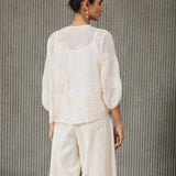 A woman stands with her back to the camera wearing a cream-colored two-piece linen set consisting of a sheer embroidered long-sleeve top and matching wide-leg trousers, paired with white slip-on shoes, set against a textured gray ribbed wall.