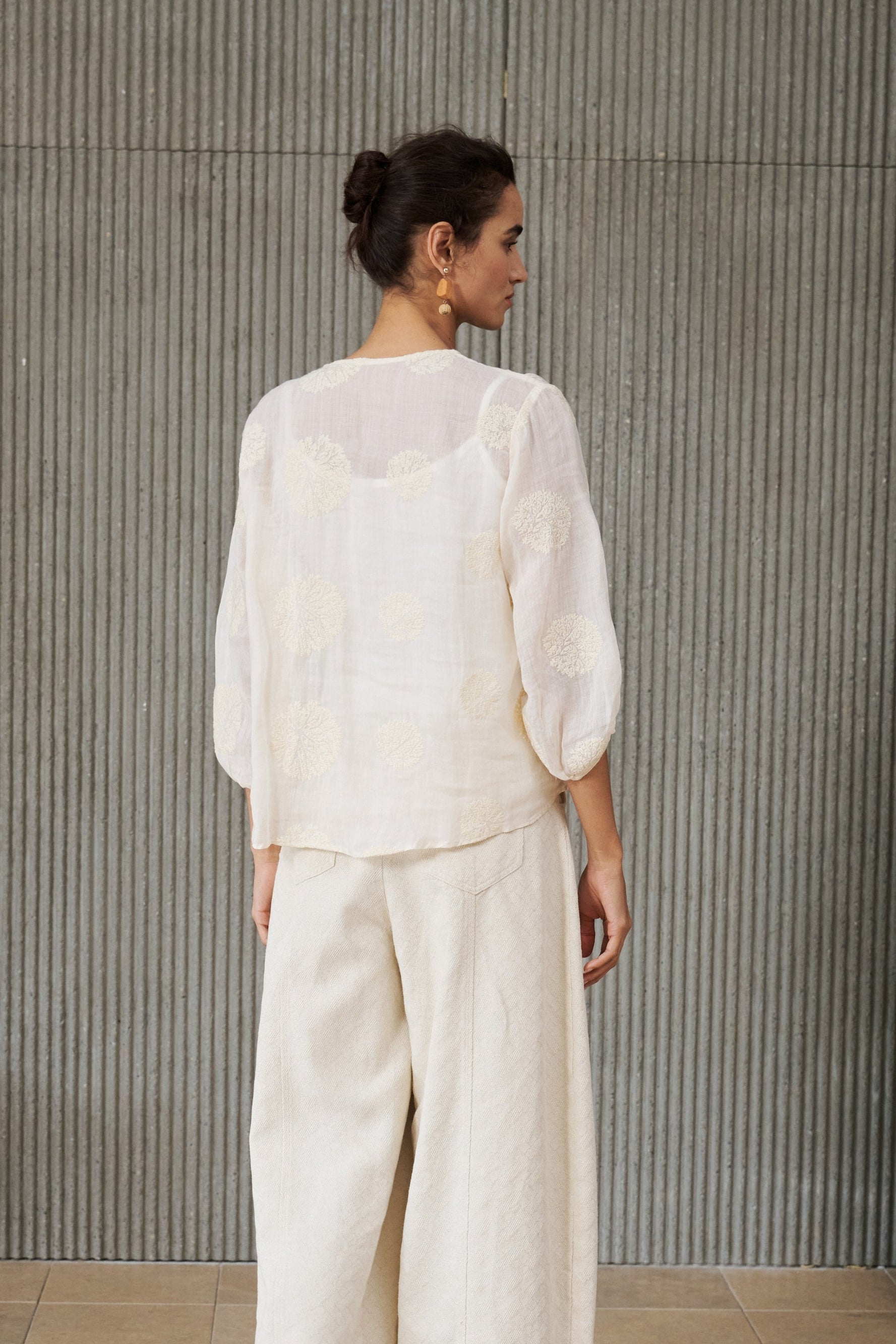 A woman stands with her back to the camera wearing a cream-colored two-piece linen set consisting of a sheer embroidered long-sleeve top and matching wide-leg trousers, paired with white slip-on shoes, set against a textured gray ribbed wall.