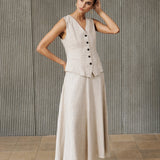 A woman models a light beige linen two-piece set consisting of a sleeveless button-front vest and a matching midi skirt, paired with white pointed-toe ankle boots, set against a textured gray ribbed wall.