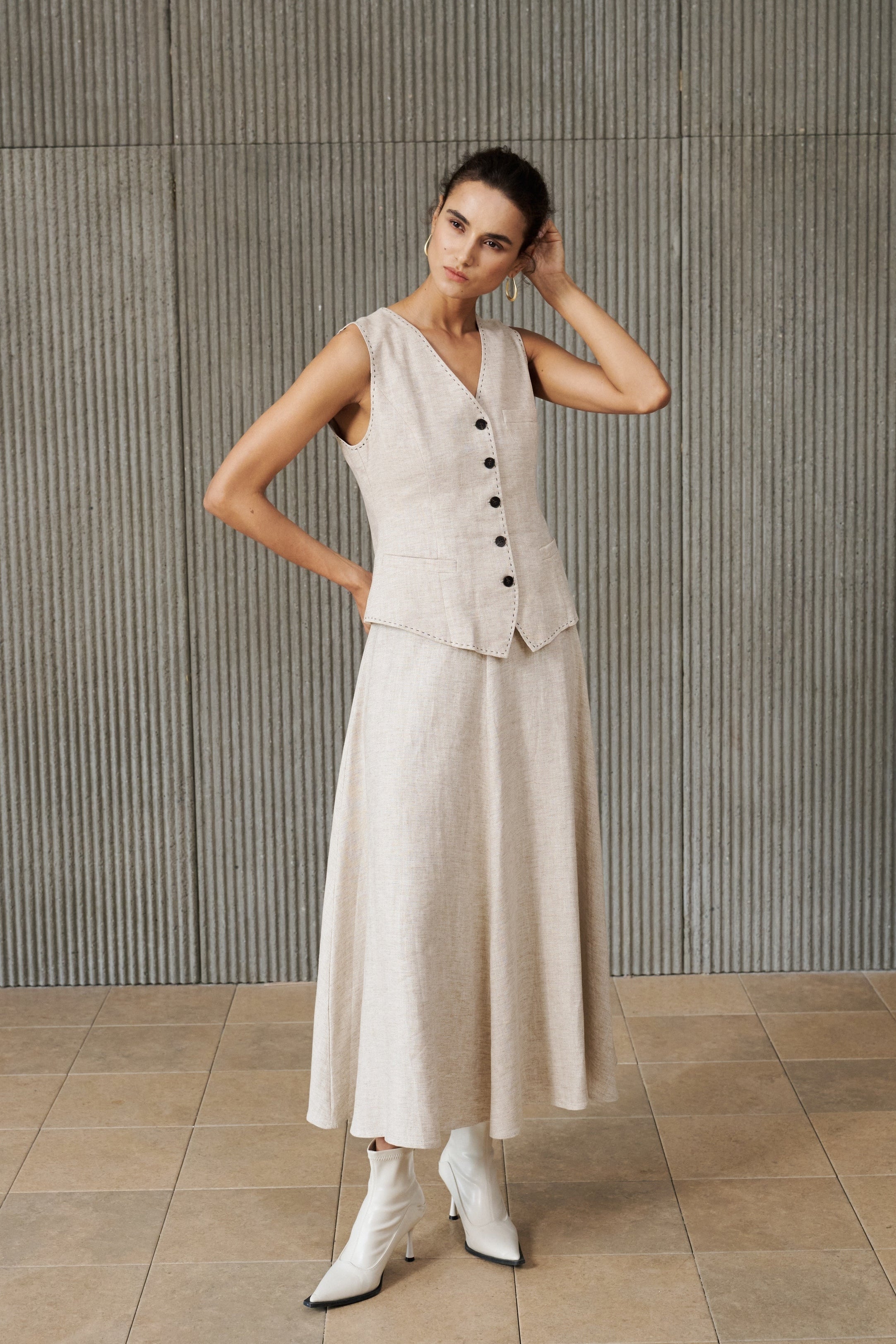 A woman models a light beige linen two-piece set consisting of a sleeveless button-front vest and a matching midi skirt, paired with white pointed-toe ankle boots, set against a textured gray ribbed wall.