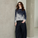 A woman with glasses stands in a minimalist studio wearing a navy and white gradient knit sweater and dark wide-leg jeans.