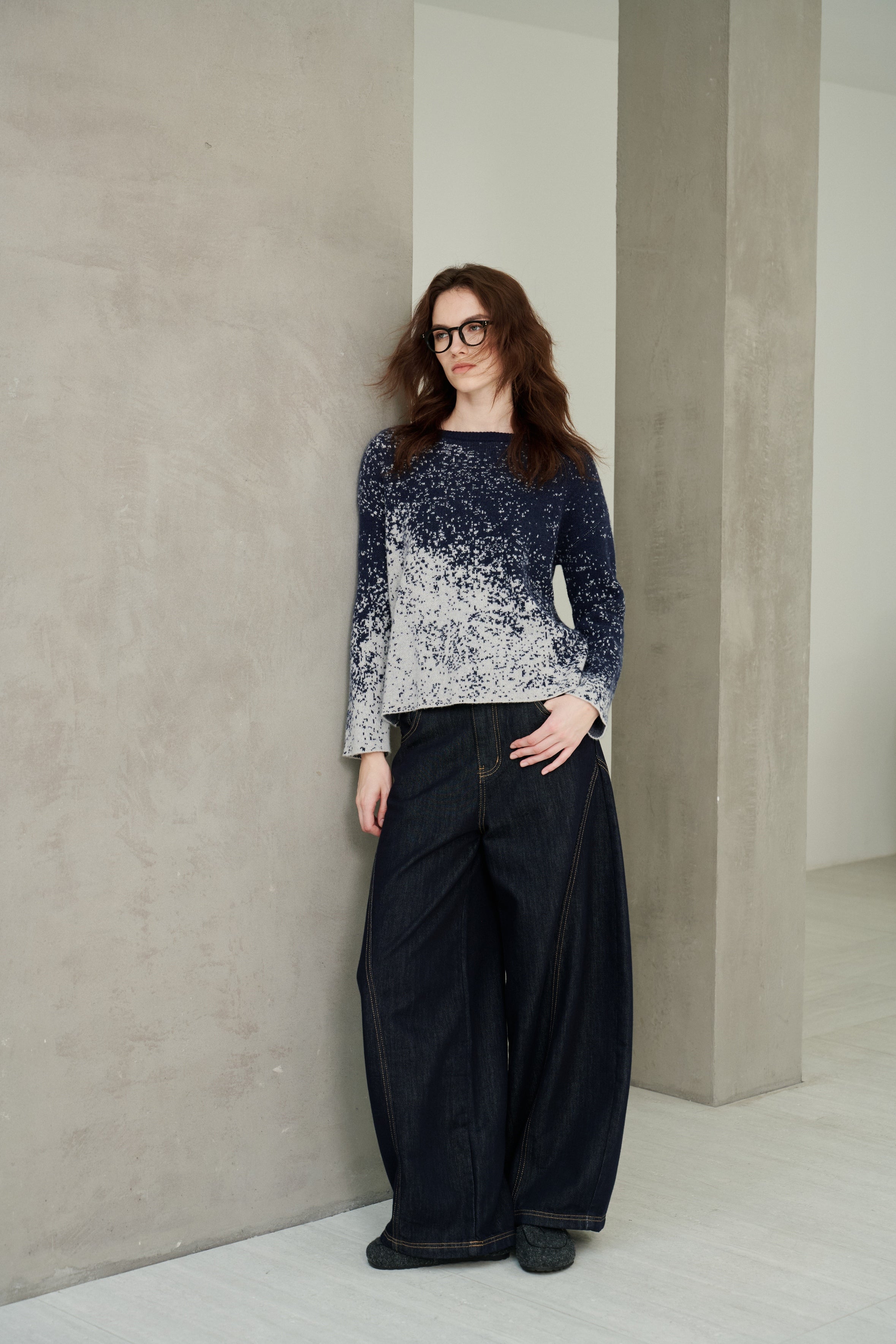 A woman with glasses stands in a minimalist studio wearing a navy and white gradient knit sweater and dark wide-leg jeans.