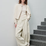 A woman stands on gray stone stairs against a light gray wall, wearing a coordinated light beige outfit with a cropped textured jacket, ribbed top, wide-leg pants, and white pointed shoes.