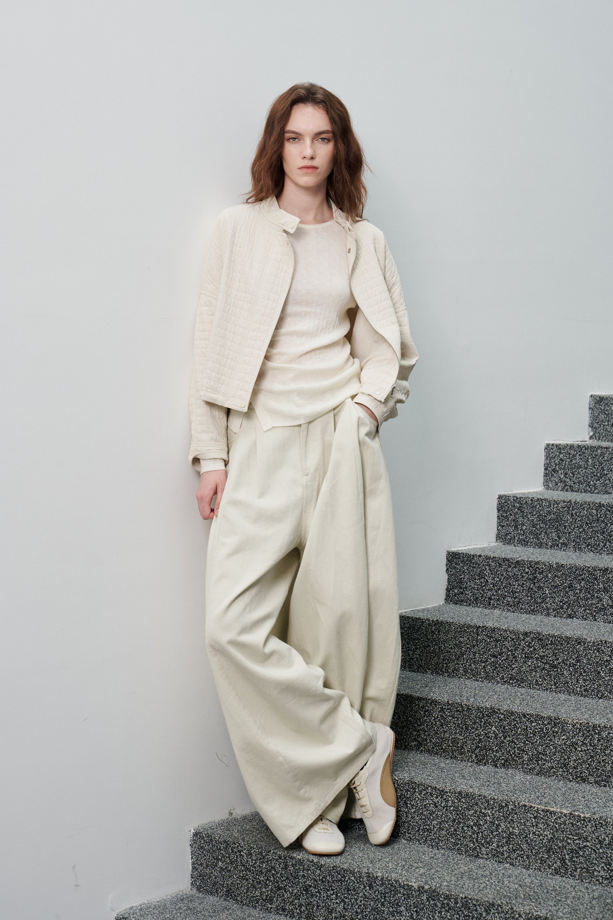 A woman stands on gray stone stairs against a light gray wall, wearing a coordinated light beige outfit with a cropped textured jacket, ribbed top, wide-leg pants, and white pointed shoes.