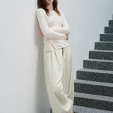 A woman wearing a cream-colored long-sleeve knit top and matching wide-leg trousers stands on a gray stone staircase against a white wall.