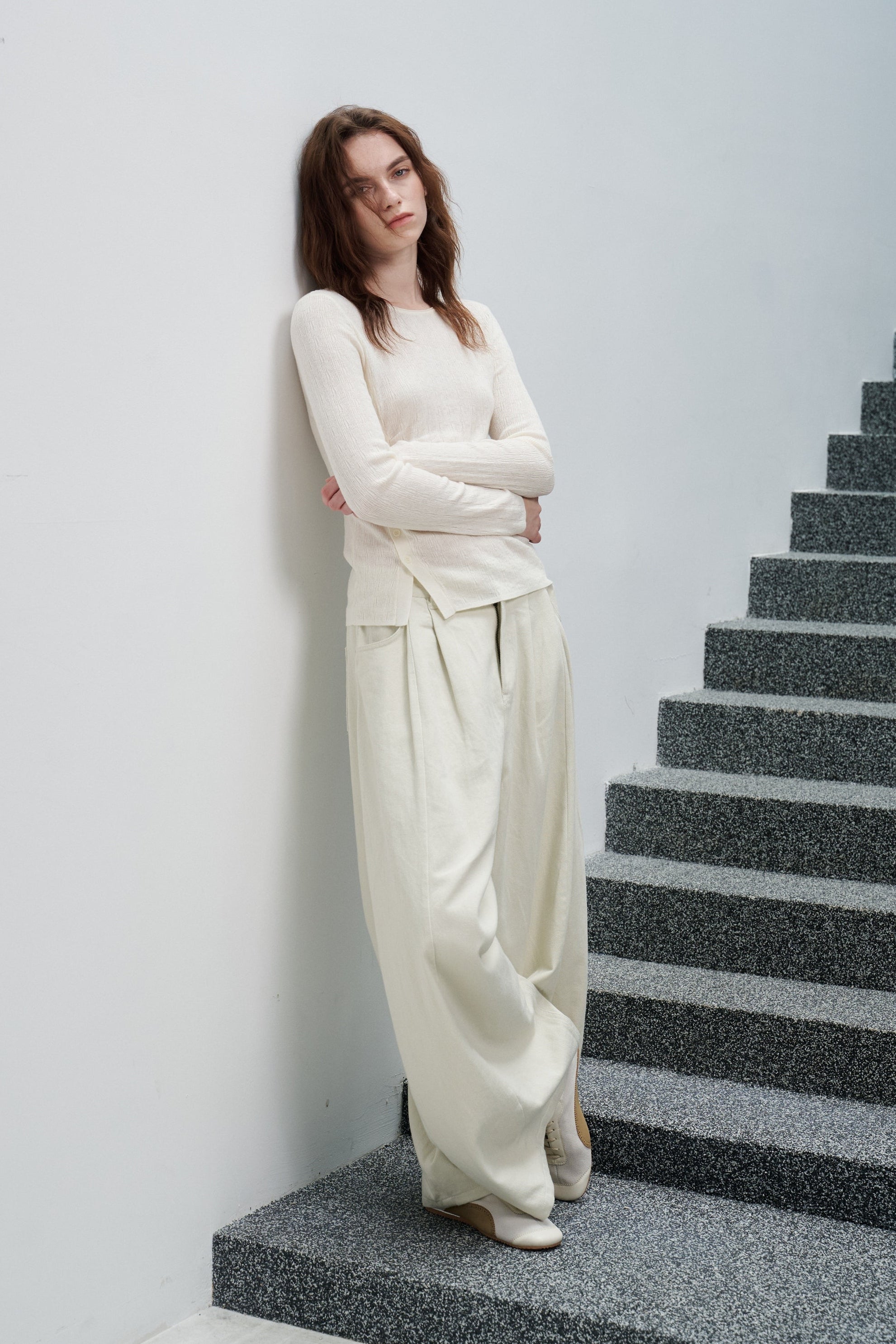 A woman wearing a cream-colored long-sleeve knit top and matching wide-leg trousers stands on a gray stone staircase against a white wall.