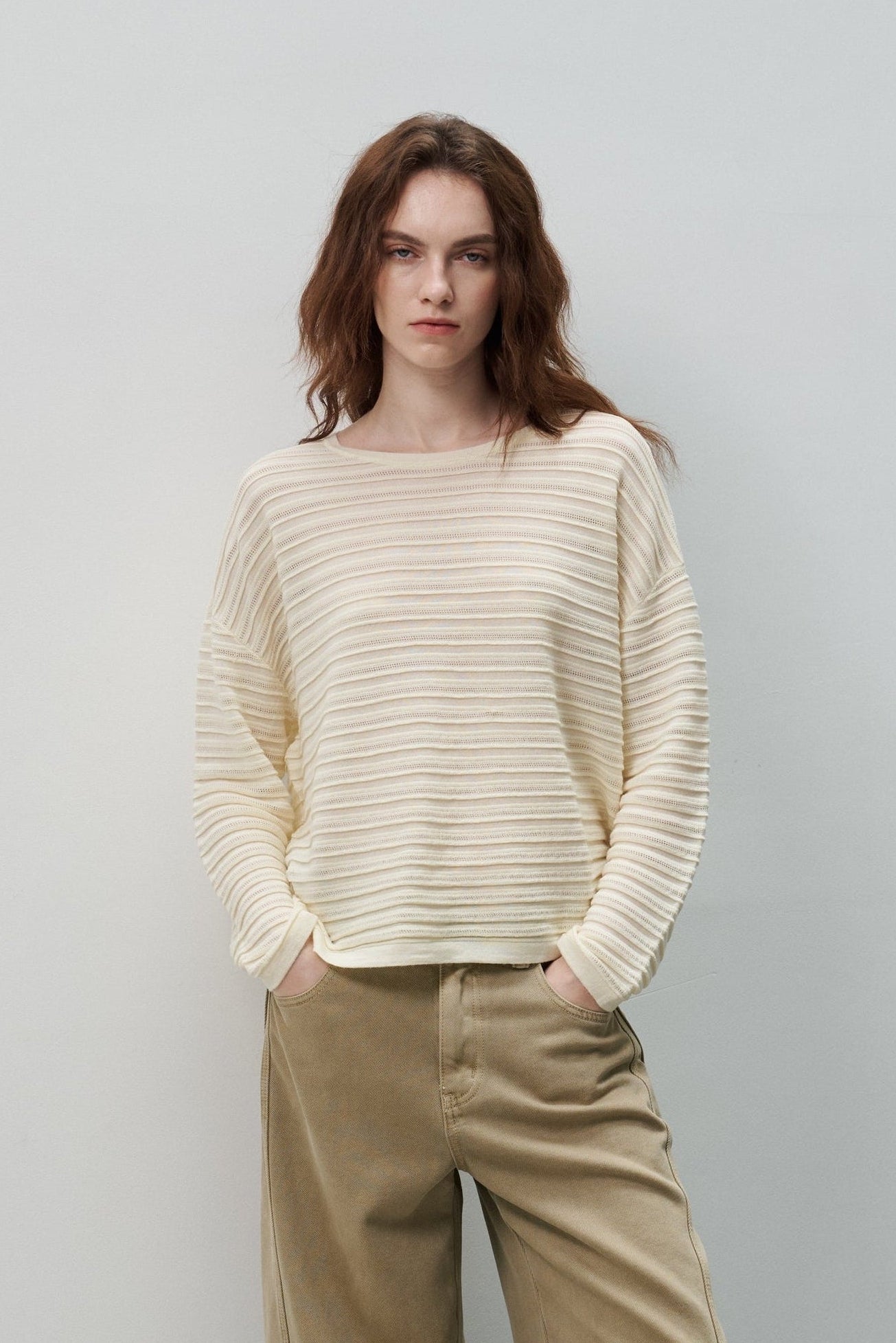 A woman stands on gray stone stairs against a light gray wall, wearing a cream ribbed knit sweater and wide-leg tan pants with white shoes.
