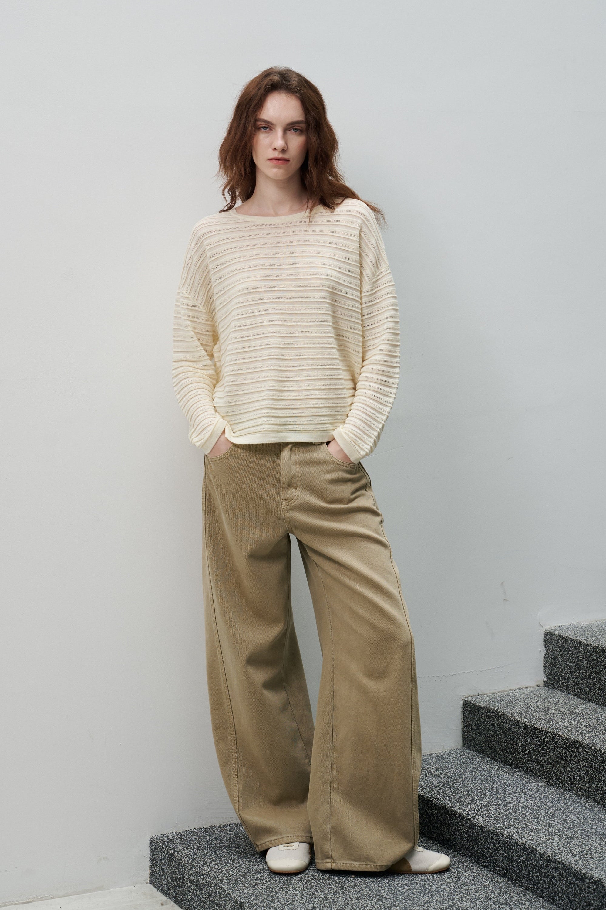 A woman stands on gray stone stairs against a light gray wall, wearing a cream ribbed knit sweater and wide-leg tan pants with white shoes.