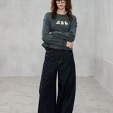 A woman wearing a dark gray crewneck sweatshirt with a small graphic on the chest and dark wide-leg jeans, standing in a minimalist gray room.