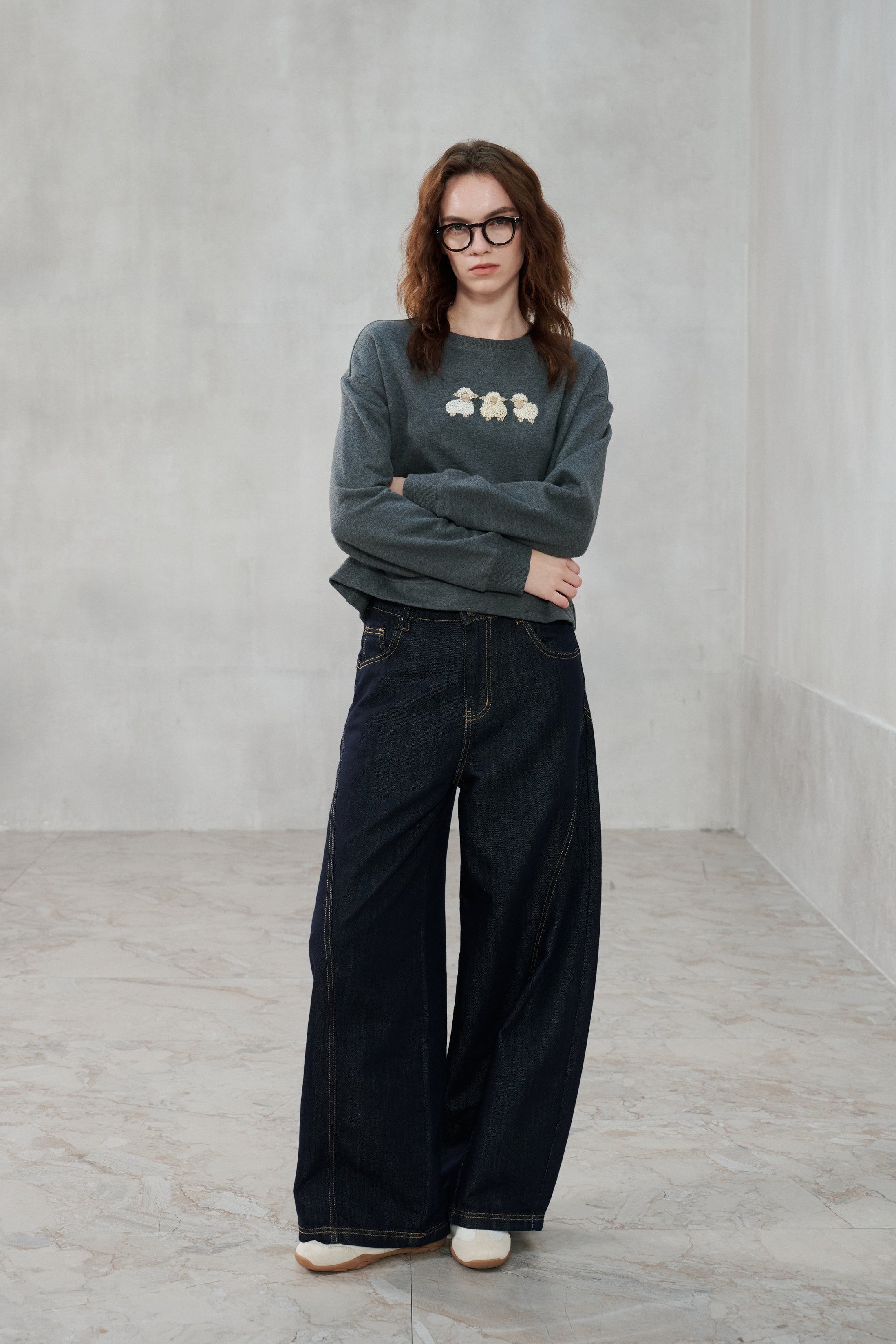 A woman wearing a dark gray crewneck sweatshirt with a small graphic on the chest and dark wide-leg jeans, standing in a minimalist gray room.