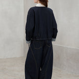 A model viewed from behind wearing a dark navy blue denim jacket and matching wide-leg denim pants, standing in a minimalist gray concrete room.