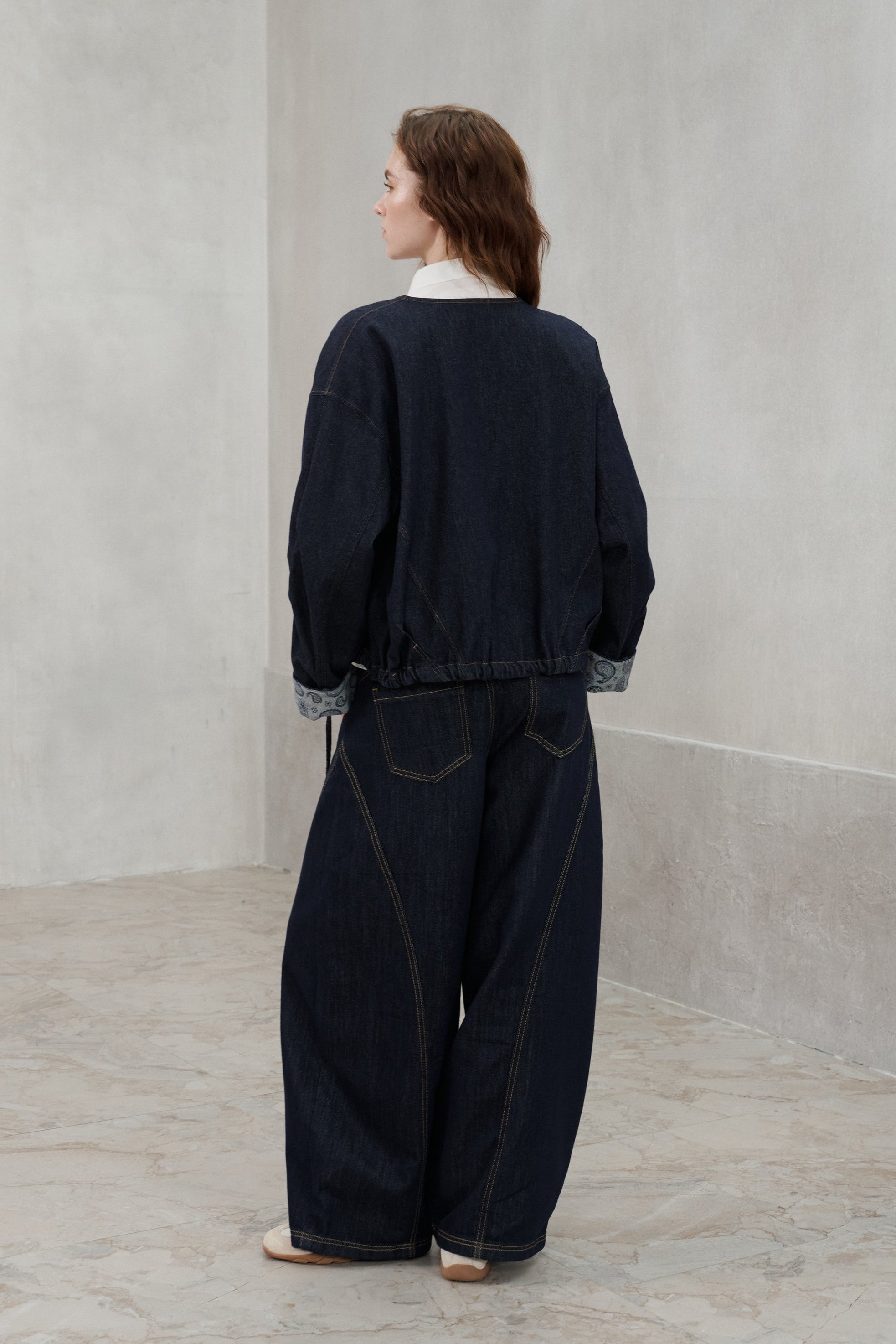 A model viewed from behind wearing a dark navy blue denim jacket and matching wide-leg denim pants, standing in a minimalist gray concrete room.