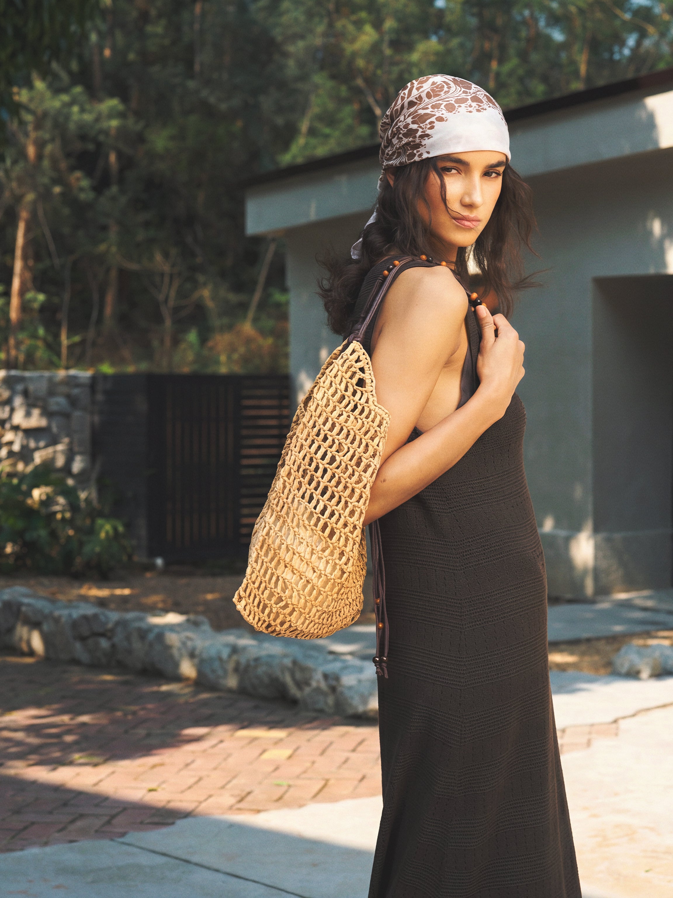 A woman wearing a black sleeveless maxi dress and a patterned headscarf stands outdoors in front of a modern concrete building, carrying a large woven straw tote bag.