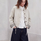 A model stands in a minimalist concrete room wearing a light beige cropped jacket over a white shirt, paired with dark wide-leg jeans and white slip-on shoes.