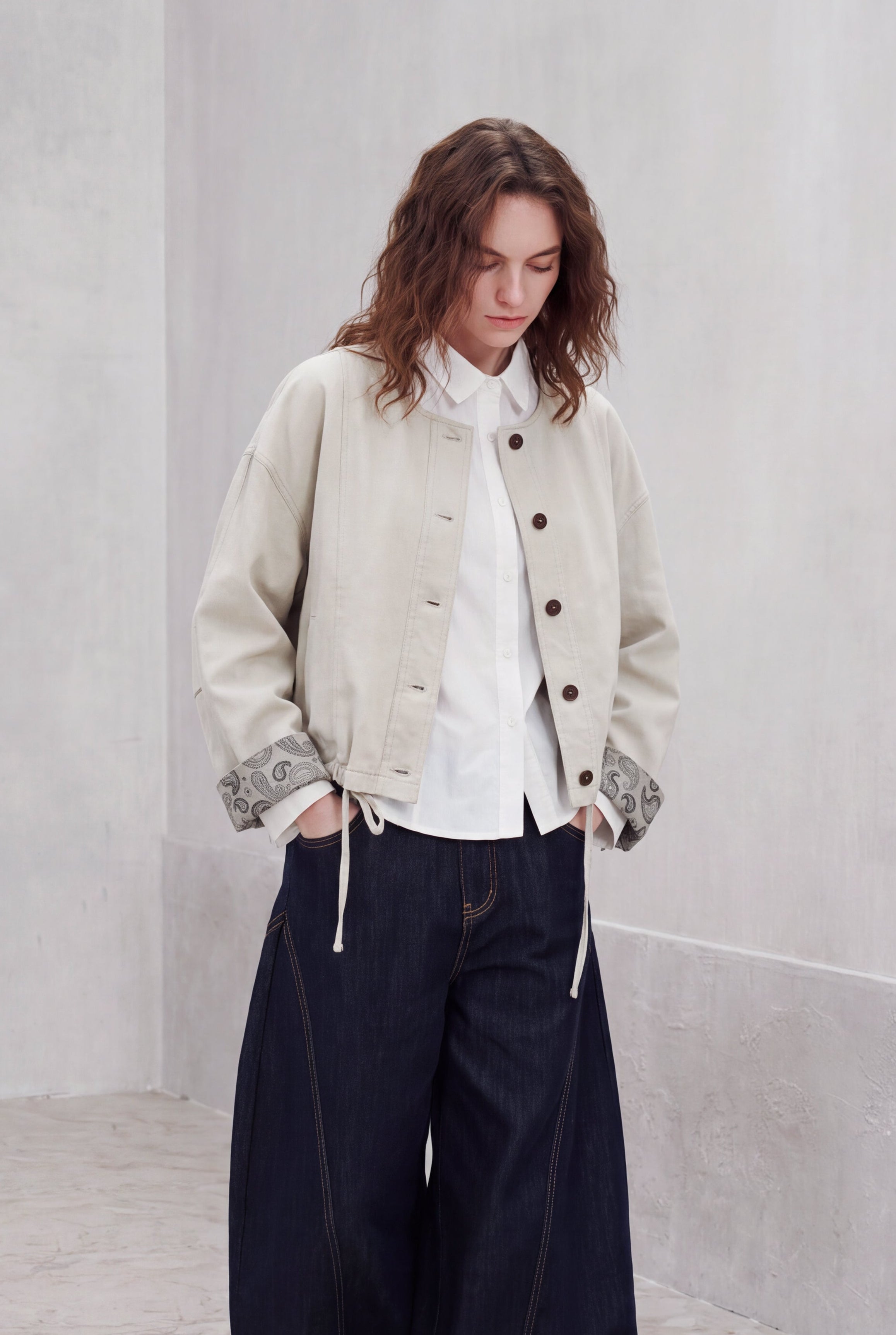 A model stands in a minimalist concrete room wearing a light beige cropped jacket over a white shirt, paired with dark wide-leg jeans and white slip-on shoes.