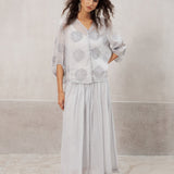 A woman modeling a light gray two-piece set consisting of a short-sleeve button-up blouse with circular embroidery and a matching long pleated skirt, standing against a textured light gray wall.