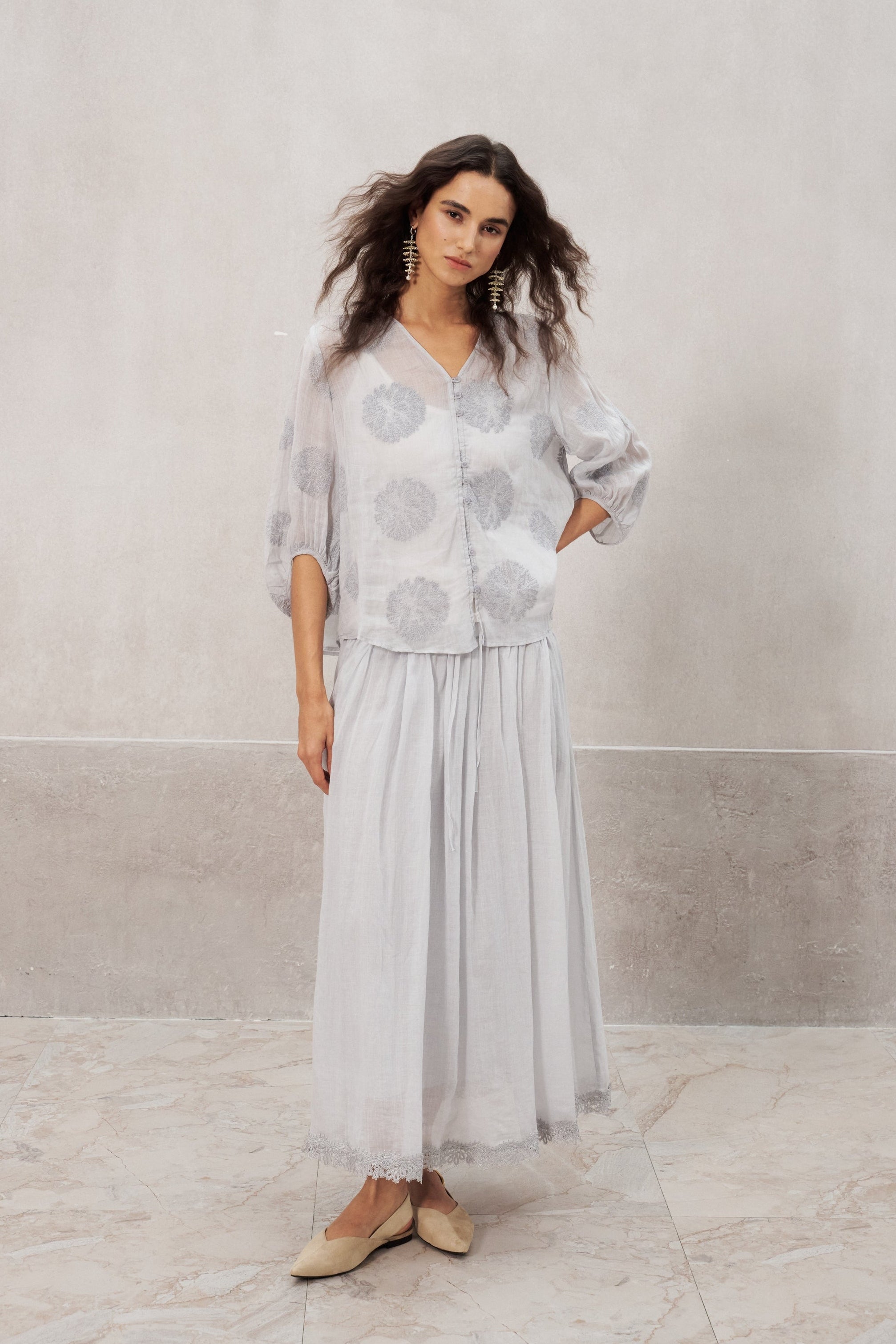A woman modeling a light gray two-piece set consisting of a short-sleeve button-up blouse with circular embroidery and a matching long pleated skirt, standing against a textured light gray wall.