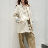 A woman stands on gray stone steps against a white wall, wearing a cream oversized jacket over a beige striped top and beige wide-leg pants.