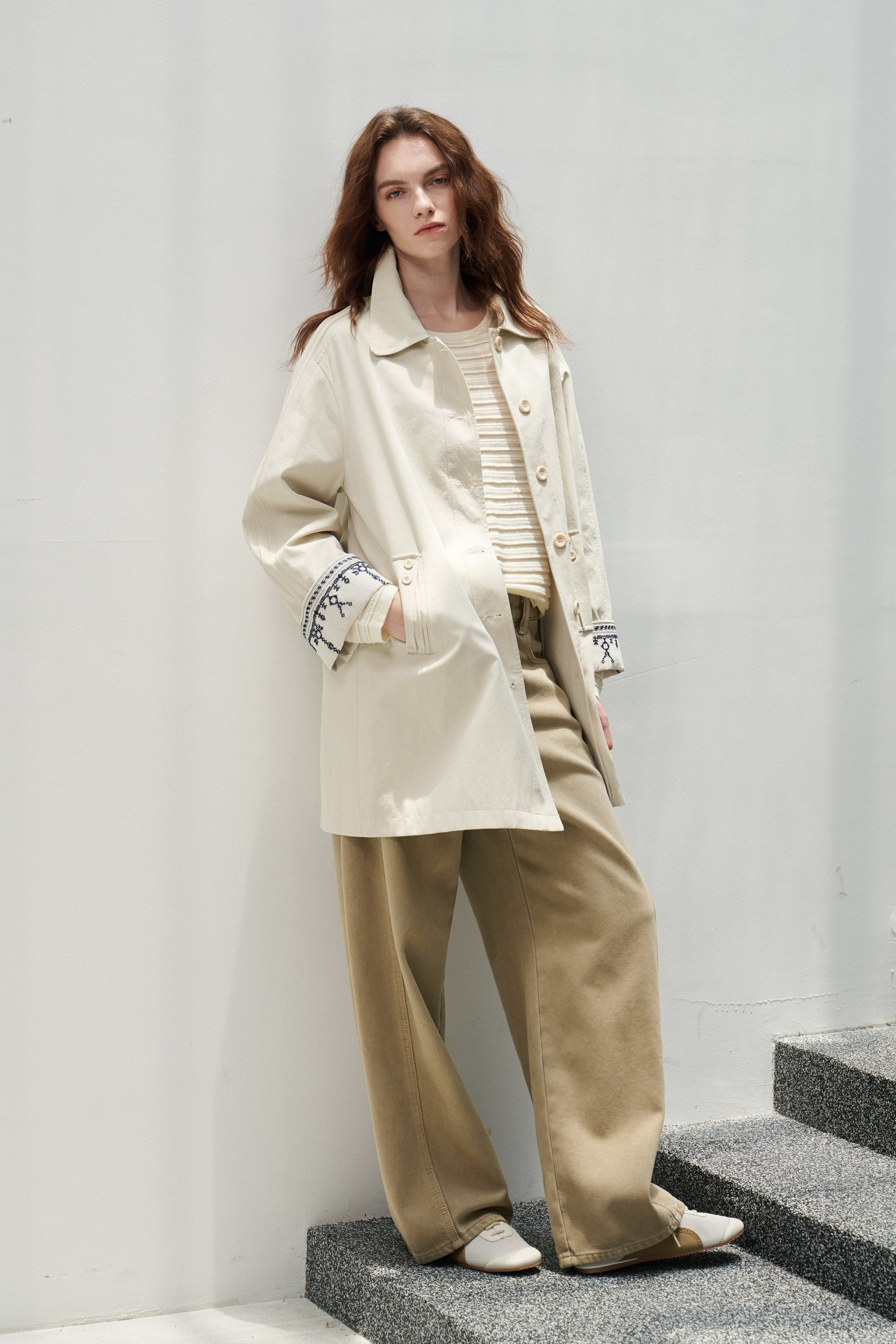 A woman stands on gray stone steps against a white wall, wearing a cream oversized jacket over a beige striped top and beige wide-leg pants.